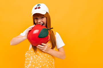 red-haired girl with a red handbag in the shape of an apple is grimacing at the camera on a yellow background