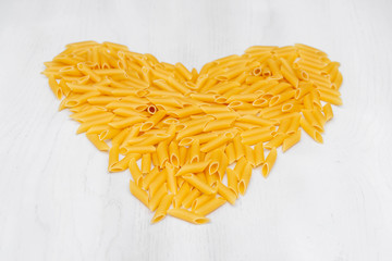Pasta penne rigate in the shape of a heart on a white background wooden table. Top view, copy space