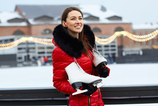 Young Woman In Red Winter Overall Combs Stands With Smile Happy Face At Open Air Outdoor Ice Rink Arena Board Fence With Wireless Earphones In Ear And Holds By Hand Hanging Skates On Shoulders