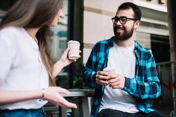 Smiling asian male laughing during conversation with caucasian female colleague during coffee break, happy hipsters friends talking to each other holding takeaway cup and telling funny stories