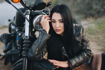 Plakat Seductive brunette girl with long hair in a black leather jacket sits near a modern motorcycle on a background of nature. Closeup portrait of a sexy woman near an expensive black bike.