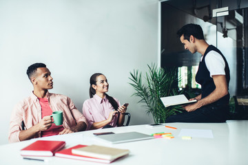 Multicultural male and female colleagues enjoying time for collaborating in coworking space with modern interior, happy entrepreneurs smiling at brainstorming meeting sitting at desktop and talking