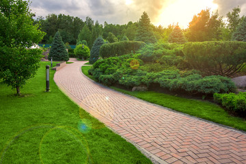 a winding pedestrian walkway made of red paving slabs with a gray stone curb in a park with a green lawn and bushes on evening with sun flare, a ground street light was installed on the lawn.
