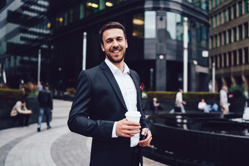 Half length portrait of happy successful male entrepreneur dressed in formal elegant wear holding takeaway cup with caffeine beverage and smiling at camera during coffee break in financial district