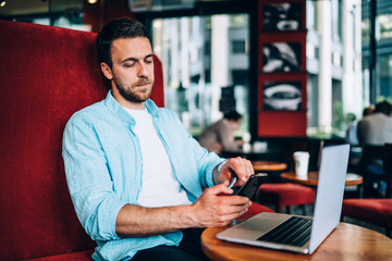 Serious skilled man in casual wear updating notification and screen settings on cellphone while working remotely with laptop device at public cafeteria, concept of technology and messaging