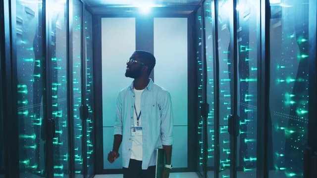 Young IT engineer walking in cyberspace room inspecting server rack cabinets. Happy professional black administrator working at secure storage data center.