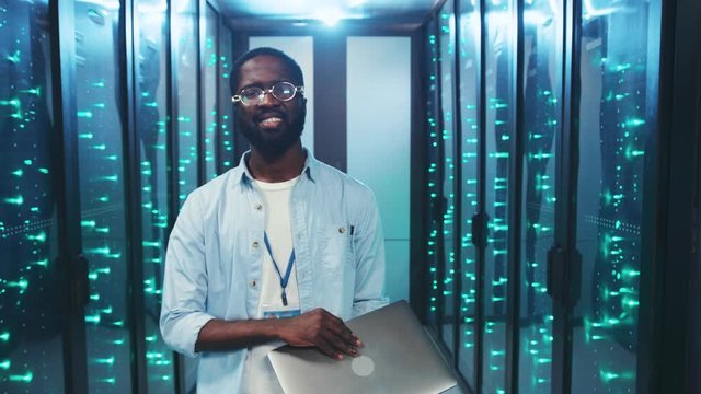 Attractive black IT technician inspecting a high secure server room smiling opening a laptop enjoying working at contemporary data center.