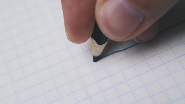 young man draws straight line using dark blue pencil on checkered paper sheet extreme close view