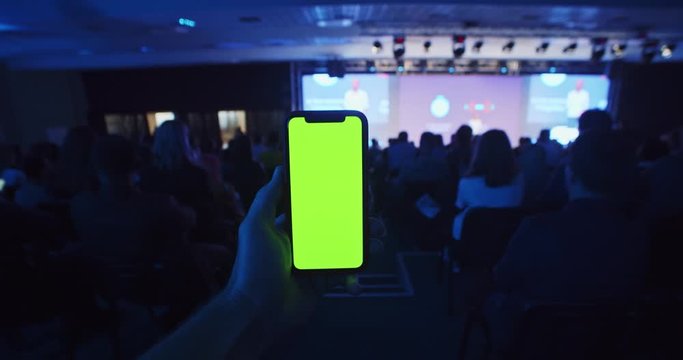 Male hand using modern smartphone with empty chroma key display in conference hall with business people and speaker on background. Training seminar. Mockup green screen. Educating lifestyle.Technology