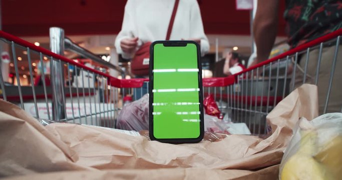 Mock-up smartphone with greenscreen app and foodstuff inside trolley in motion. Timelapse close-up of young couple taking food at checkout point shooping in supermarket.