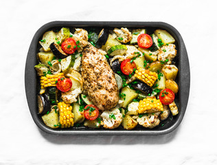 Chicken breast baked with potatoes, cauliflower, corn, eggplant, zucchini, cherry tomatoes on a baking sheet on a light background, top view. Healthy food concept
