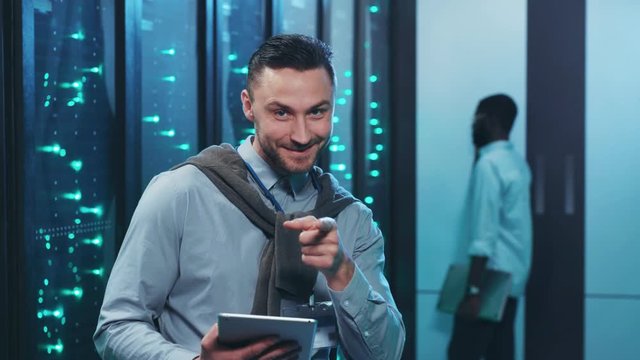 Positive IT engineer holding digital tablet pointing at camera having fun smiling coworking in secure server rack room of data center.