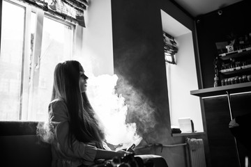 Vaping teenager. Young pretty white girl smoking an electronic cigarette opposite the window  in vape bar. Bad habit. Black and white.