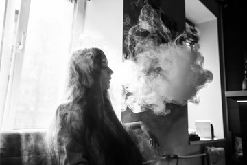 Vaping teenager. Young pretty white girl smoking an electronic cigarette opposite the window  in vape bar. Bad habit. Black and white.