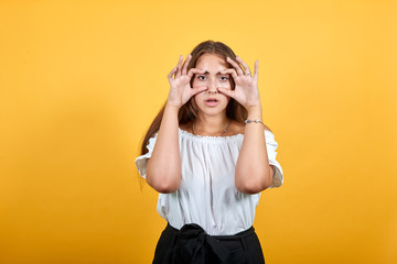 Attractive caucasian young woman in blue denim shirt doing binoculars gesture isolated on orange background in studio. People sincere emotions, lifestyle concept.