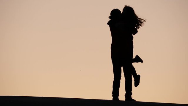 Couple happy to meet, cheerful woman rush to man, they hug and turn around. Telephoto view, silhouetted shot against clear light yellow sunset sky