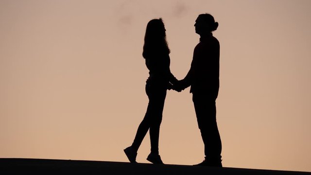 Young woman come to man, pair join hands and kiss lightly, silhouetted shot at top. Dim yellow sky on backdrop, telephoto perspective.