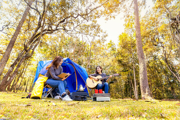 Two Beautiful young woman with guitar and tent of outdoor. Asian woman camping and backpack travel sitting relax alone in forest. A young female relaxing in front of a tent.