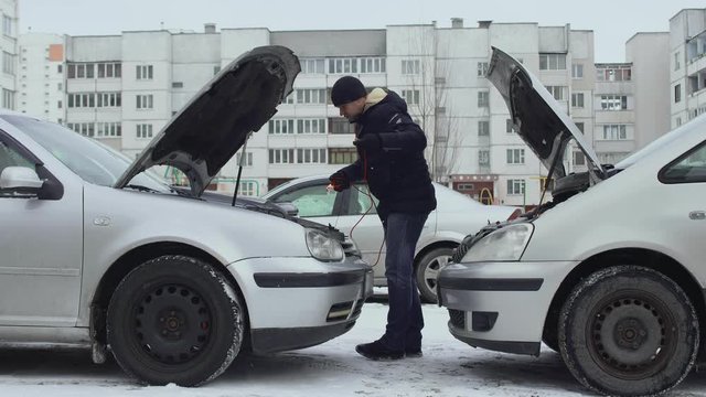 Man connects jumper clamp cables from discharged automotive battery to battery post terminal. Hoods raised up in winter daytime in parking lot of yard. Malfunction or problem with auto. Part 3