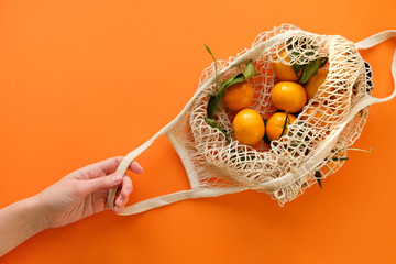 A human hand holds a string bag with tangerines on bold orange color background.