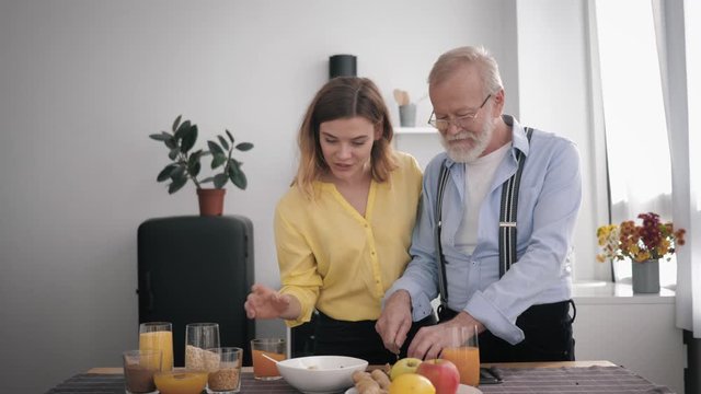 family relationship, happy peppy old man prepares a delicious breakfast in the kitchen with healthy products, mixes with his cheerful beautiful adult granddaughter at the table background of the kit