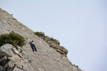A man wearing a helmet, with equipment, climbs a cliff. Via Ferrata in the south of France. Bushes, grey rocks, a blue sky.