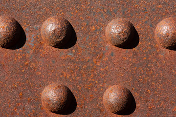 Six sunny rusty bolts head of a steel footbridge and their shadow on a rusty beam. Centered in the image.
