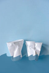 Two smashed white plastic coffee cups on a light blue background with hand drawn transparant waves on top. Zero waste, plastic free, stop sea pollution, ecological concept. Photo with copy space.