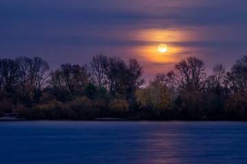 Fototapeta na wymiar Real full moon over the autumn forest close to Dnieper river in Kiev, Ukraine. Soft clouds in the dark sky cover partially our natural satellite. The water looks like a glossy blue mirror.