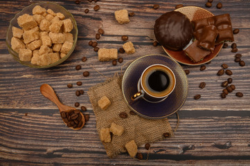 A Cup of coffee, coffee beans, brown sugar in a sugar bowl, chocolate pieces, marshmallows in chocolate on a wooden background.