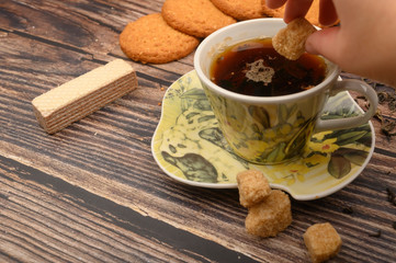 The girl's hand puts a piece of brown sugar in a mug of black tea, oatmeal cookies, tea leaves, waffles, brown sugar on a wooden background. Close up.