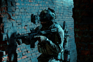 Fototapeta premium Man in uniform with machine gun and night-vision device move inside broken building. Airsoft soldier in night building