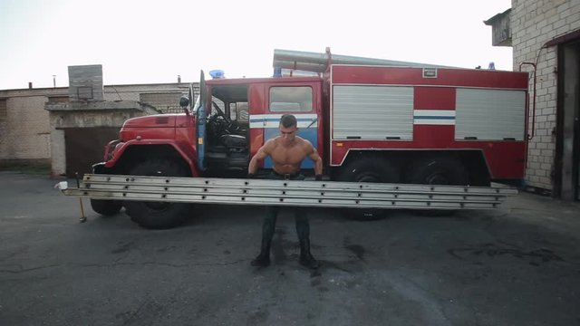 Handsome young man doing exercises with a metal ladder in front of a fire engine