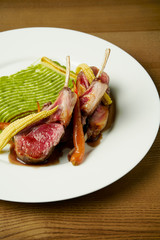 Juicy rack of a young lamb with vegetables and green pasta on a white plate. Restaurant serve from the chef. Wooden background. Close up, selective focus. Food flat lay