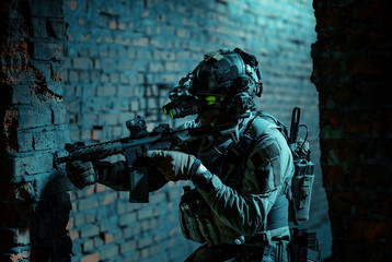 Man in uniform with machine gun and turned on night vision device move between brick wall. Closeup airsoft soldier with green light on face in night