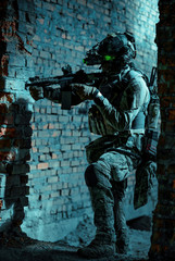 Fototapeta premium Man in uniform with machine gun and turned on night vision device move between walls Airsoft soldier with green light on face in night. Side view