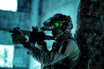 Man in uniform with machine gun and turned on night vision device beside brick wall. Airsoft...