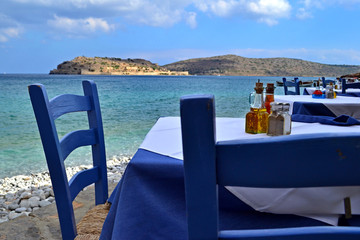 Cozy tables of street cafe in the shade. Tables with tablecloths. On the table - salt shaker, vessels with olive oil and vinegar. All in Mediterranean style. Against the blue sea and Spinalonga island