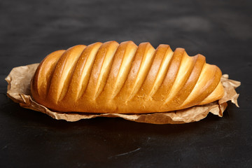 Loaf, white wheat bread on baking paper on black background, side view. Baked whole bun on dark stone table
