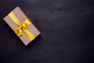 Craft paper gift box with golden, yellow ribbon bow on black wooden background. Copy space.