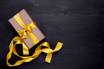 Craft paper gift box with golden, yellow ribbon bow on black wooden background. Copy space. Gold ribbon.