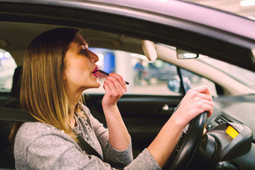 Young woman sitting in the car on the driver's seat putting the make up on her lips using pen crayon looking to the mirror while waiting on the parking at night - Powered by Adobe
