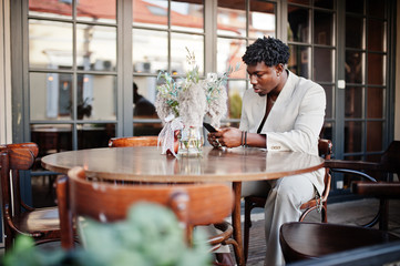 Stylish afro man in beige old school suit sitting on cafe with mobile phone. Fashionable young African male in casual jacket on bare torso.