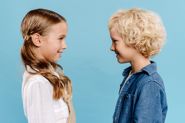 side view of smiling and cute kids looking at each other isolated on blue