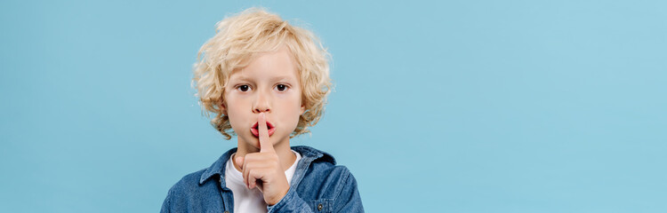 panoramic shot of cute kid showing secret gesture and looking at camera isolated on blue - obrazy, fototapety, plakaty