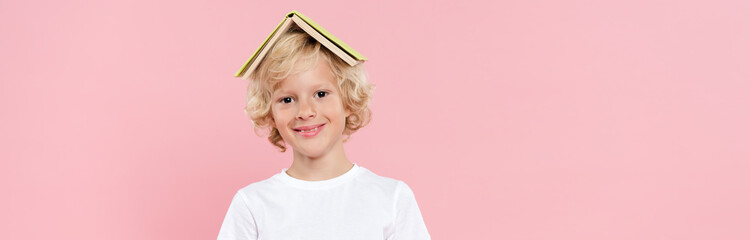 panoramic shot of smiling kid with book on head isolated on pink