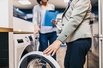 Saleswoman working in appliances store and showing to senior woman washing machines.