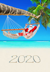Santa Claus relax in hammock at tropical palm beach with handwritten caption of New Year 2020 on white sand, Christmas vacation concept