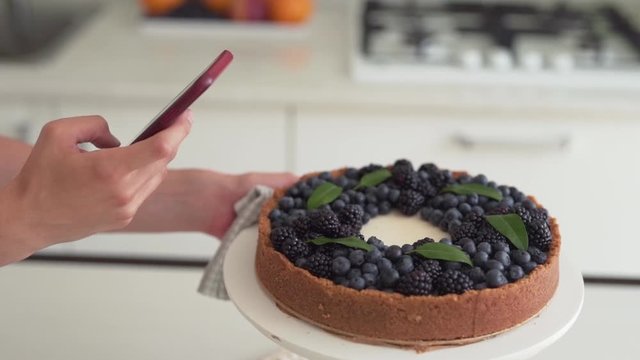 Close-up scene of a female cook who has just made a cheesecake and now has taking a photo of it on a phone.