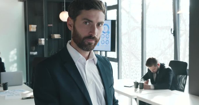 Young Attractive Businessman Posing on Camera in Modern Office, Wearing Formal Clothes. Looking Happy and Smiling while Looking at Camera. Business People working in the Background.